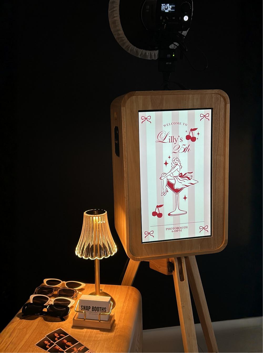Digital photo booth with screen displaying a design, surrounded by props on a dark background.